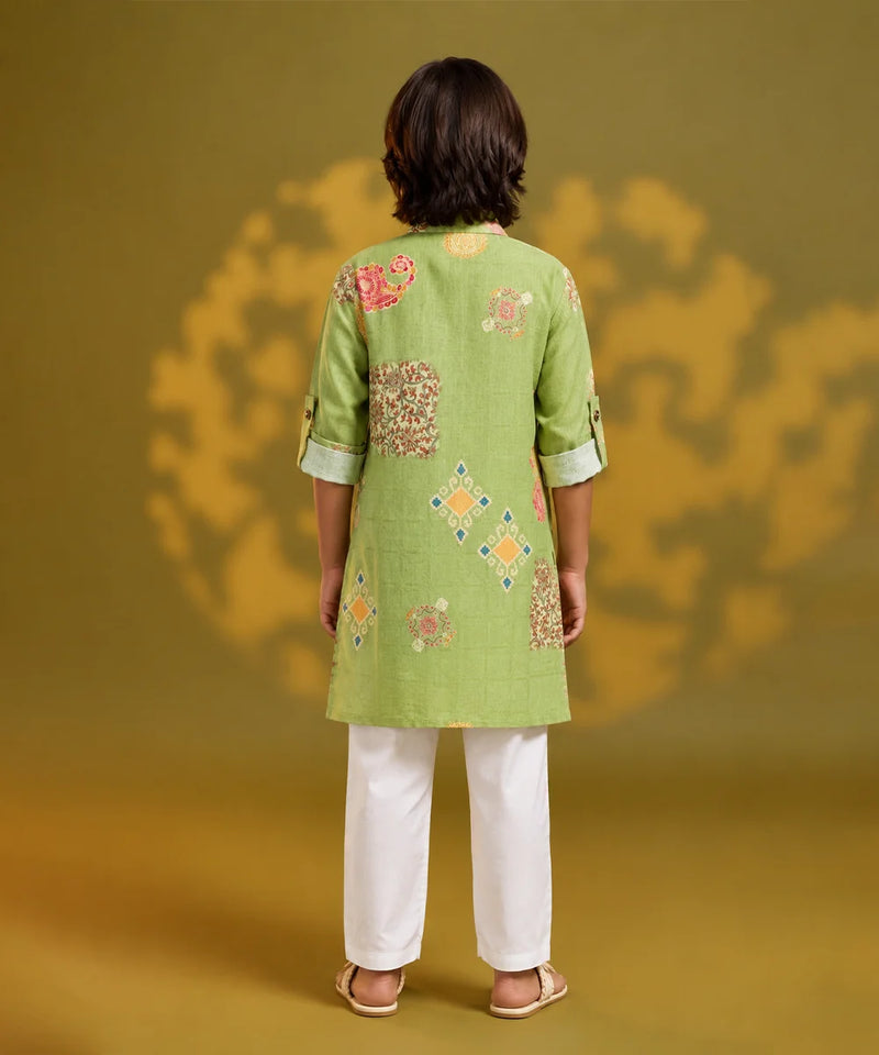A model viewed from the back wearing a light green kurta with colorful floral and geometric embroidery, paired with white trousers, set against a warm yellow background with soft shadow details.