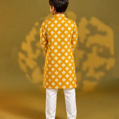 A child viewed from behind wearing a mustard yellow kurta with a white geometric diamond pattern, paired with white trousers and sandals, standing against a warm yellow background with soft shadow details.