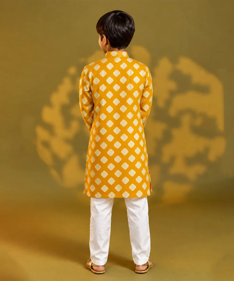 A child viewed from behind wearing a mustard yellow kurta with a white geometric diamond pattern, paired with white trousers and sandals, standing against a warm yellow background with soft shadow details.