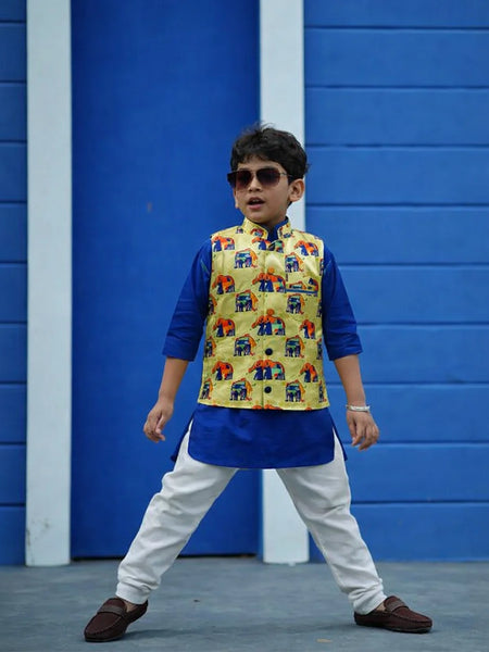 Child wearing a colorful vest over a blue kurta with white pants against a blue wall.