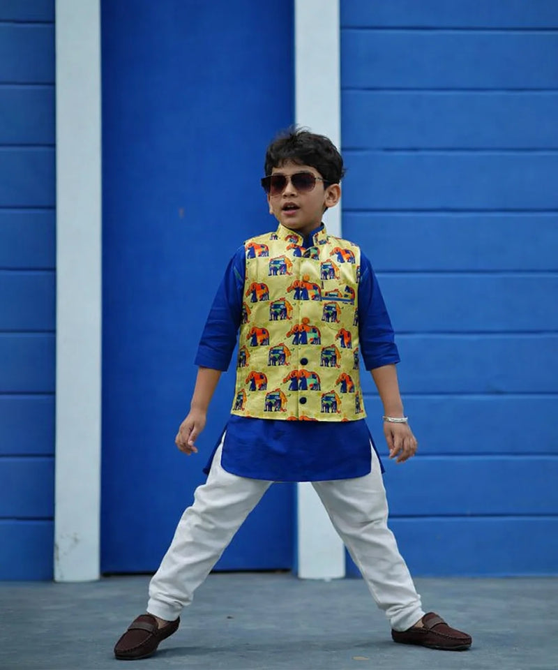 Child wearing a colorful vest over a blue kurta with white pants against a blue wall.