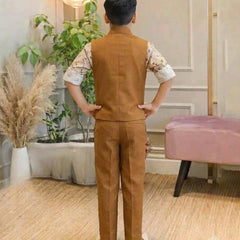 A child viewed from behind wearing a brown vest and matching trousers over a white patterned short-sleeve shirt, standing in an indoor room with plants, a lamp, and a pink bench.