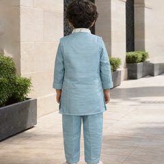 Child in a light blue suit standing in an outdoor setting with stone walls and plants.