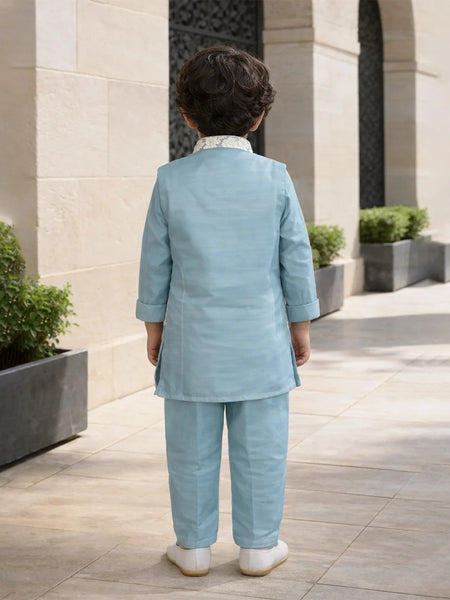 Child in a light blue suit standing in an outdoor setting with stone walls and plants.