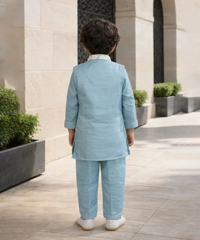 Child in a light blue suit standing in an outdoor setting with stone walls and plants.