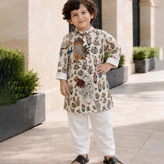 A young boy wearing a cream-colored kurta with a detailed multicolor floral and bird print, paired with white trousers and black slide sandals, standing outdoors in front of a beige stone building.