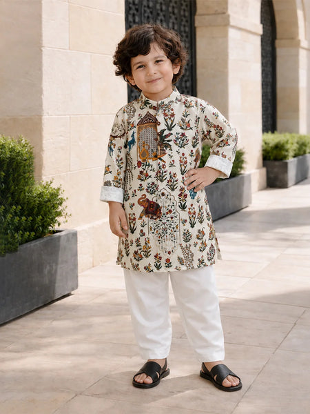 A young boy wearing a cream-colored kurta with a detailed multicolor floral and bird print, paired with white trousers and black slide sandals, standing outdoors in front of a beige stone building.
