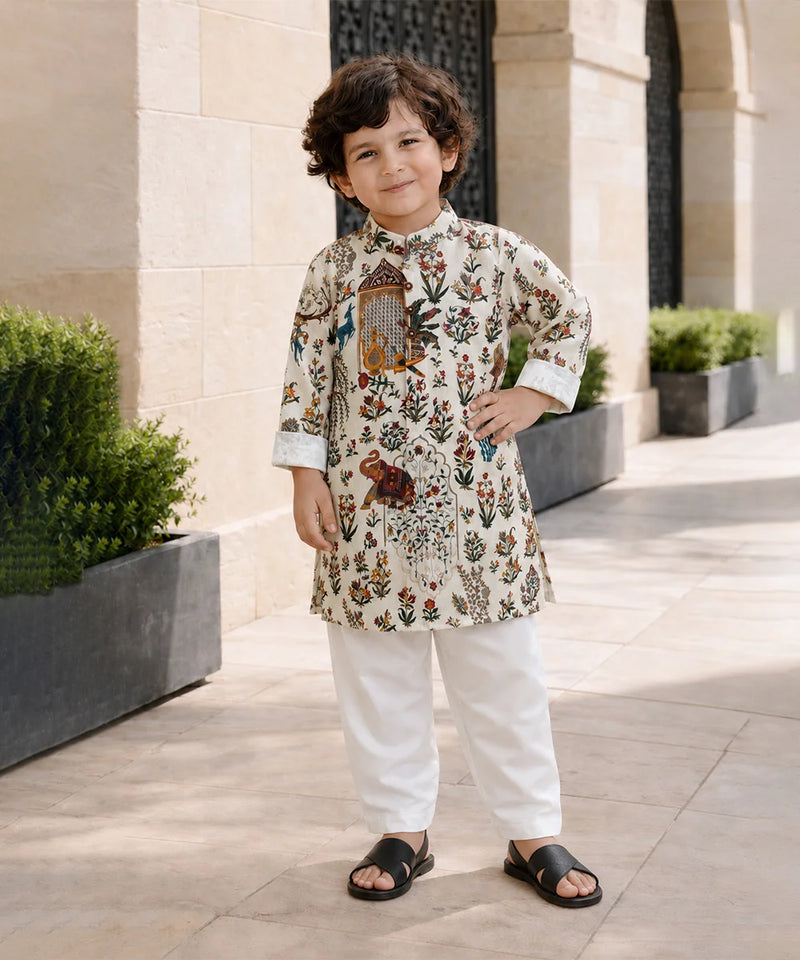 A young boy wearing a cream-colored kurta with a detailed multicolor floral and bird print, paired with white trousers and black slide sandals, standing outdoors in front of a beige stone building.