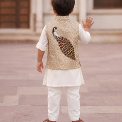 A toddler boy seen from behind wearing a white traditional outfit with a gold sequined vest featuring a large embroidered peacock design on the back, standing in an outdoor architectural setting.