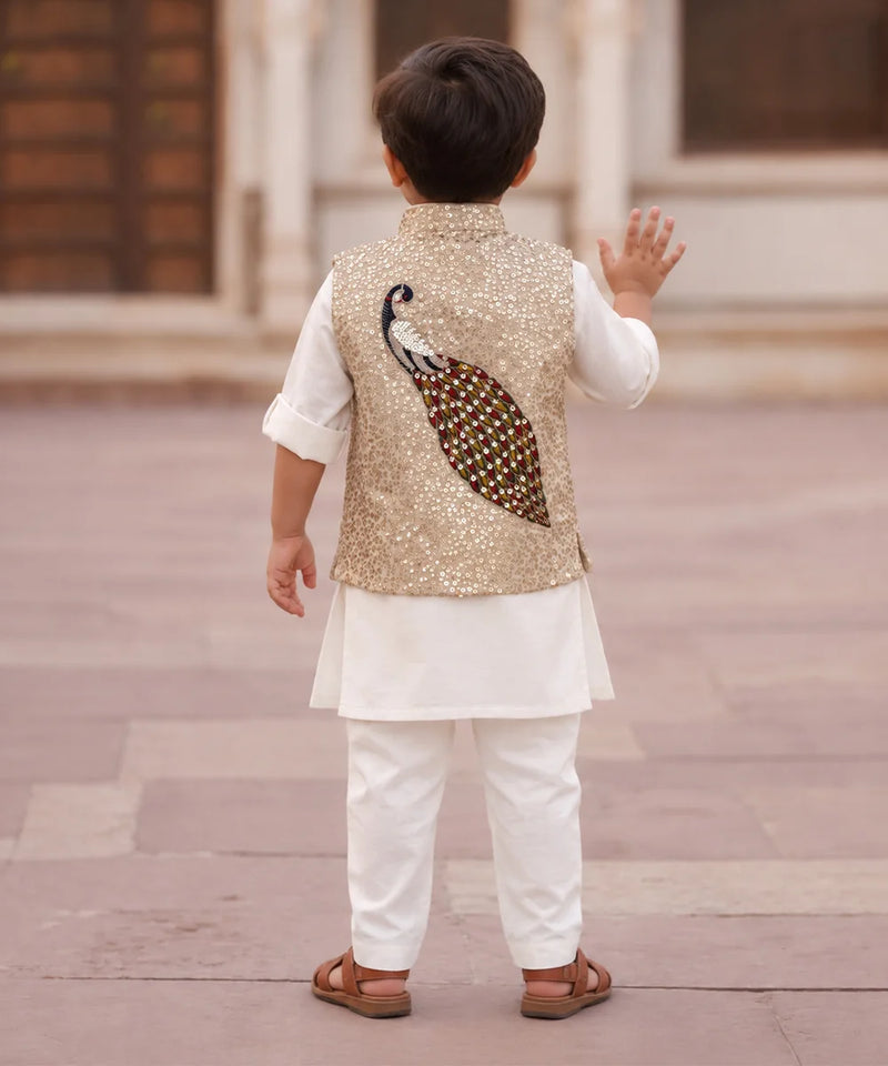 A toddler boy seen from behind wearing a white traditional outfit with a gold sequined vest featuring a large embroidered peacock design on the back, standing in an outdoor architectural setting.