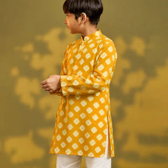 A child viewed from behind wearing a mustard yellow kurta with a white geometric diamond pattern, paired with white trousers and sandals, standing against a warm yellow background with soft shadow details.