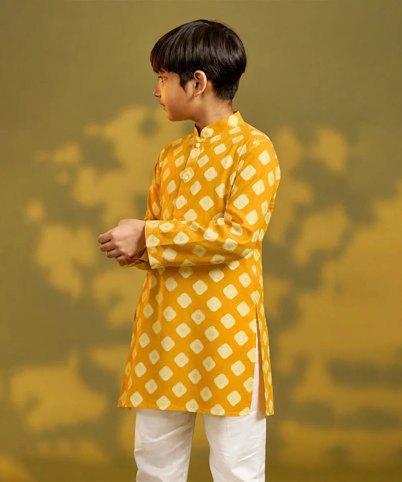 A child viewed from behind wearing a mustard yellow kurta with a white geometric diamond pattern, paired with white trousers and sandals, standing against a warm yellow background with soft shadow details.