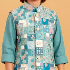 A child wearing a teal embroidered vest with a patchwork floral design over a matching teal long-sleeve shirt, set against a solid beige background.