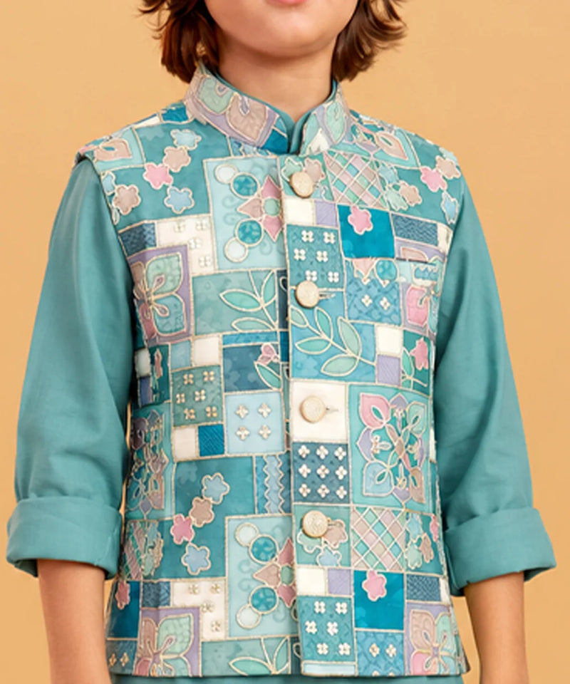 A child wearing a teal embroidered vest with a patchwork floral design over a matching teal long-sleeve shirt, set against a solid beige background.