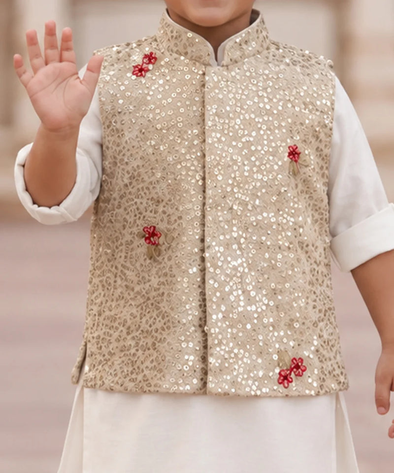 A child wearing a beige sequined vest with red floral embroidery over a white kurta, with one hand raised in a wave.