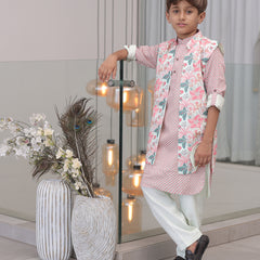 A young boy wearing a traditional Indian kurta pajama set with a floral print, consisting of a kurta and pyjama along with a long waistcoat, standing indoors with decorative lights and vases in the background.