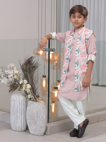 A young boy wearing a traditional Indian kurta pajama set with a floral print, consisting of a kurta and pyjama along with a long waistcoat, standing indoors with decorative lights and vases in the background.