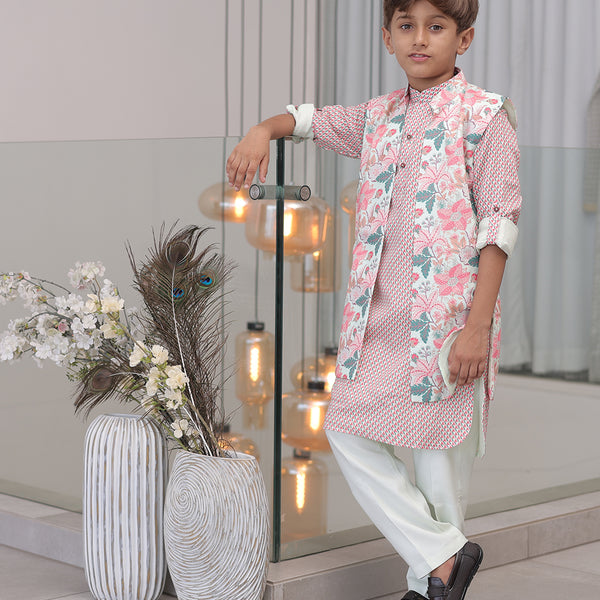 A young boy wearing a traditional Indian kurta pajama set with a floral print, consisting of a kurta and pyjama along with a long waistcoat, standing indoors with decorative lights and vases in the background.