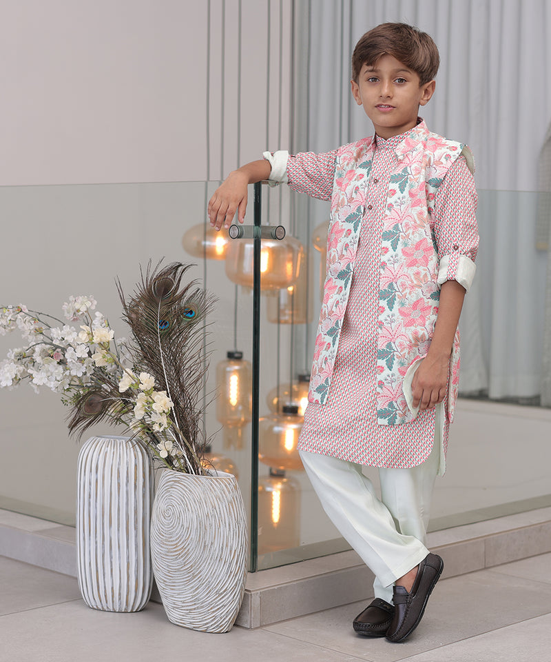 A young boy wearing a traditional Indian kurta pajama set with a floral print, consisting of a kurta and pyjama along with a long waistcoat, standing indoors with decorative lights and vases in the background.