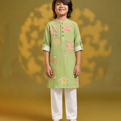 A young boy stands against a warm olive-green studio backdrop wearing a light green embroidered kurta with white pants and beige sandals. The kurta features a mandarin collar, three-quarter sleeves with rolled cuffs, and decorative floral and geometric embroidery in pink, orange, and yellow.