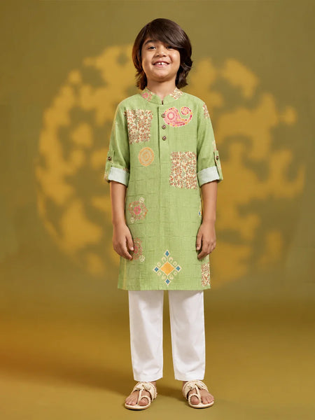 A young boy stands against a warm olive-green studio backdrop wearing a light green embroidered kurta with white pants and beige sandals. The kurta features a mandarin collar, three-quarter sleeves with rolled cuffs, and decorative floral and geometric embroidery in pink, orange, and yellow.