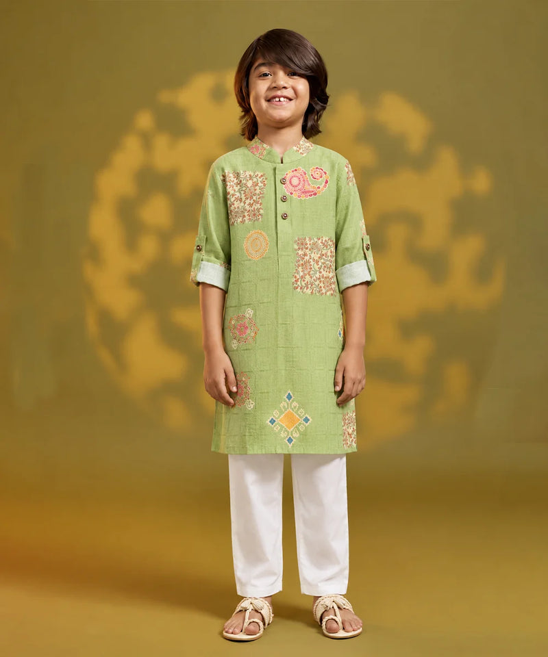 A young boy stands against a warm olive-green studio backdrop wearing a light green embroidered kurta with white pants and beige sandals. The kurta features a mandarin collar, three-quarter sleeves with rolled cuffs, and decorative floral and geometric embroidery in pink, orange, and yellow.