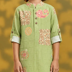 A child wearing a light green kurta with pink and orange floral and paisley patterns, shown from the shoulders to the waist against a warm yellow background.