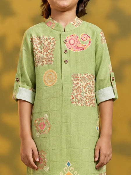 A child wearing a light green kurta with pink and orange floral and paisley patterns, shown from the shoulders to the waist against a warm yellow background.