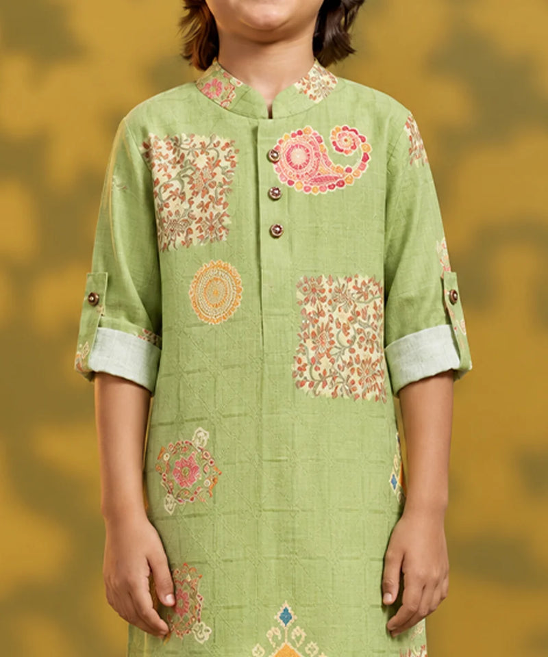 A child wearing a light green kurta with pink and orange floral and paisley patterns, shown from the shoulders to the waist against a warm yellow background.