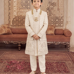 A young boy stands in a traditional Indian room wearing an off-white embroidered sherwani and cream trousers, accessorized with a gold chain necklace and beige shoes.
