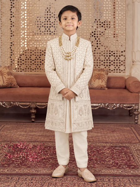 A young boy stands in a traditional Indian room wearing an off-white embroidered sherwani and cream trousers, accessorized with a gold chain necklace and beige shoes.