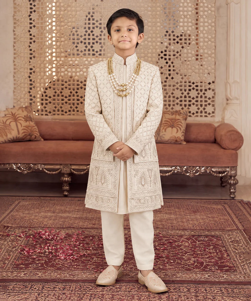 A young boy stands in a traditional Indian room wearing an off-white embroidered sherwani and cream trousers, accessorized with a gold chain necklace and beige shoes.
