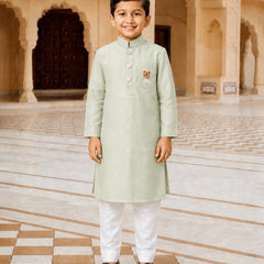 A young boy stands in a grand, arched courtyard wearing a light sage green kurta with white trousers and black loafers. The kurta features a small embroidered motif on the left chest.