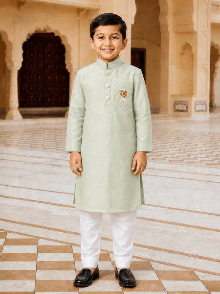 A young boy stands in a grand, arched courtyard wearing a light sage green kurta with white trousers and black loafers. The kurta features a small embroidered motif on the left chest.