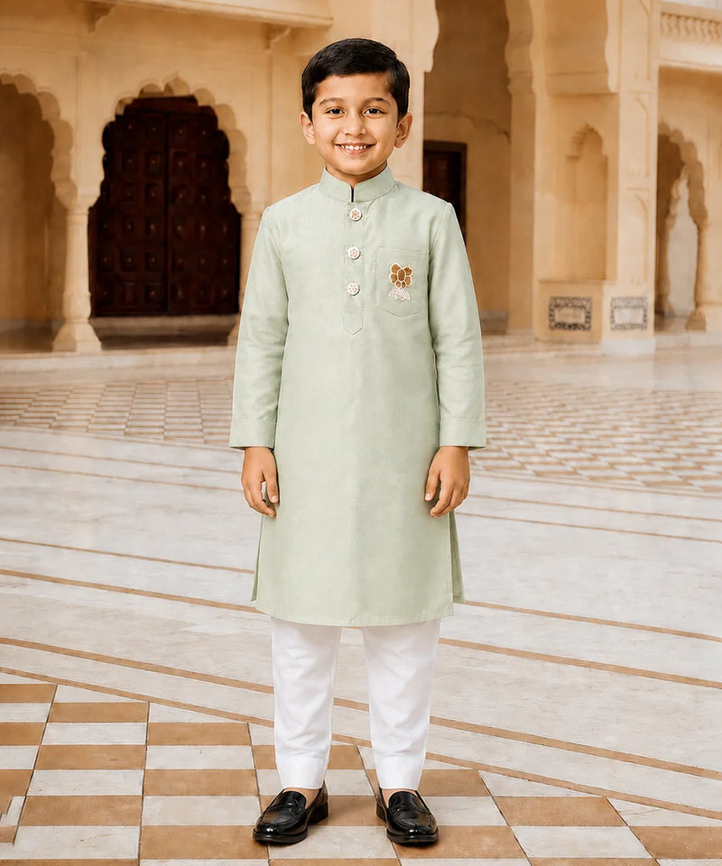 A young boy stands in a grand, arched courtyard wearing a light sage green kurta with white trousers and black loafers. The kurta features a small embroidered motif on the left chest.