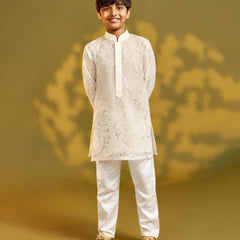A young boy modeling a traditional Indian outfit consisting of a white embroidered kurta and white trousers, standing against a warm olive-green studio backdrop.