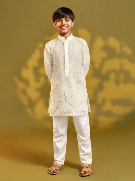 A young boy modeling a traditional Indian outfit consisting of a white embroidered kurta and white trousers, standing against a warm olive-green studio backdrop.