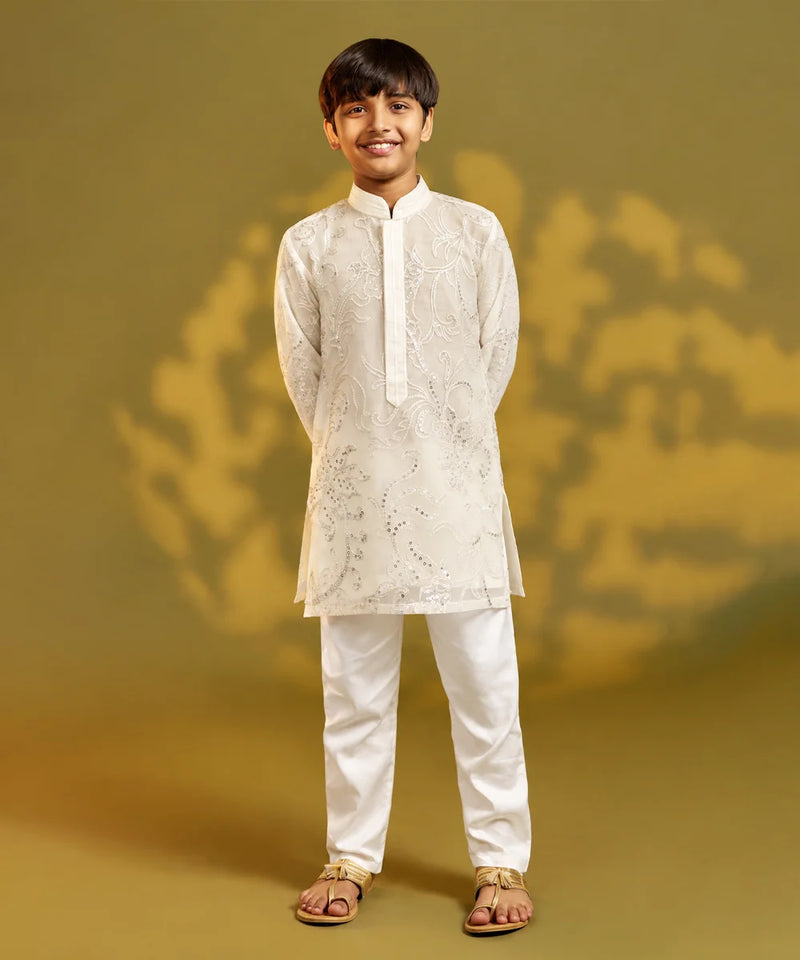 A young boy modeling a traditional Indian outfit consisting of a white embroidered kurta and white trousers, standing against a warm olive-green studio backdrop.