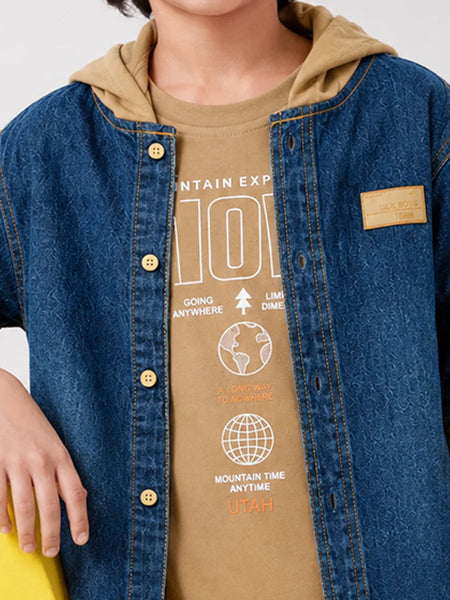 A close-up of a person wearing a blue denim overshirt over a tan graphic T-shirt with a tan hooded lining. The shirt has white and orange text and globe graphics, and a small tan patch is visible on the overshirt chest.