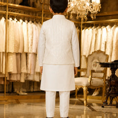 A young boy viewed from behind wearing a traditional white Indian outfit consisting of a kurta, trousers, and a matching embroidered jacket, standing in a luxury boutique with gold-framed clothing racks and a chandelier in the background.