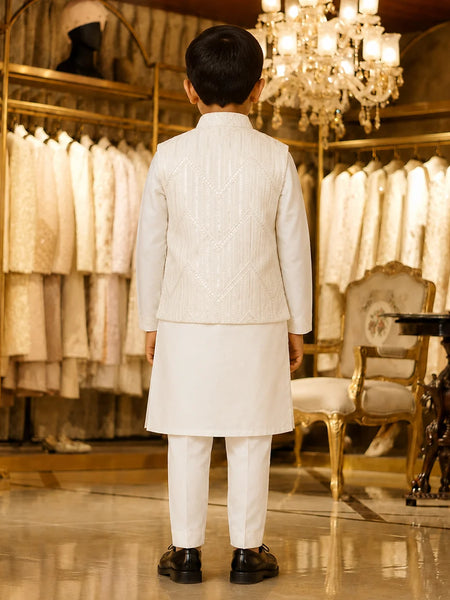 A young boy viewed from behind wearing a traditional white Indian outfit consisting of a kurta, trousers, and a matching embroidered jacket, standing in a luxury boutique with gold-framed clothing racks and a chandelier in the background.
