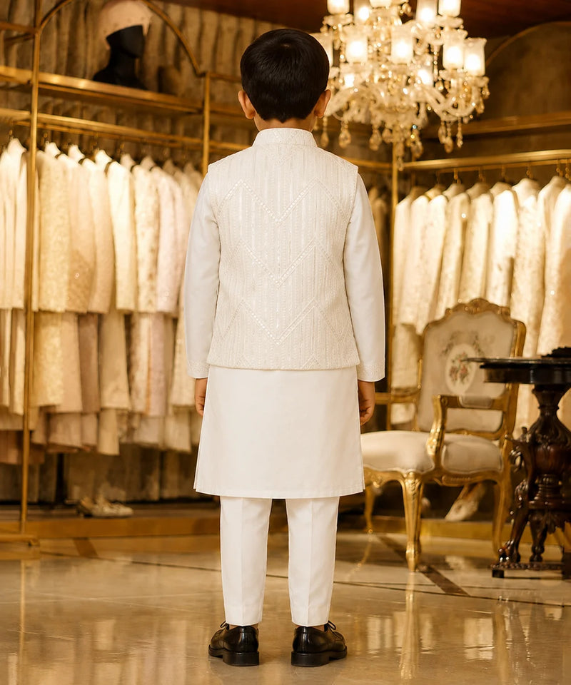 A young boy viewed from behind wearing a traditional white Indian outfit consisting of a kurta, trousers, and a matching embroidered jacket, standing in a luxury boutique with gold-framed clothing racks and a chandelier in the background.