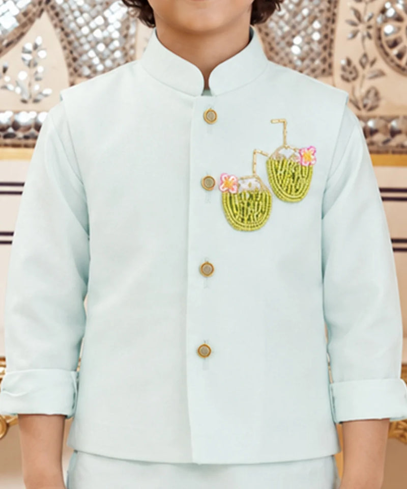 A child wearing a light mint green traditional Indian jacket with gold buttons and embroidered coconut motifs on the chest, paired with matching trousers.