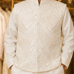 A close-up of a person wearing a white embroidered traditional jacket over a white long-sleeve shirt, with a softly blurred warm-toned indoor background.