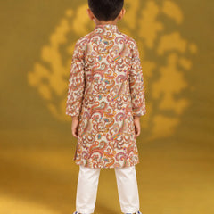 A child viewed from the back wearing a long-sleeve beige kurta with an intricate orange, red, and green paisley pattern, paired with white trousers and blue sneakers, set against a yellow background with a matching shadow.