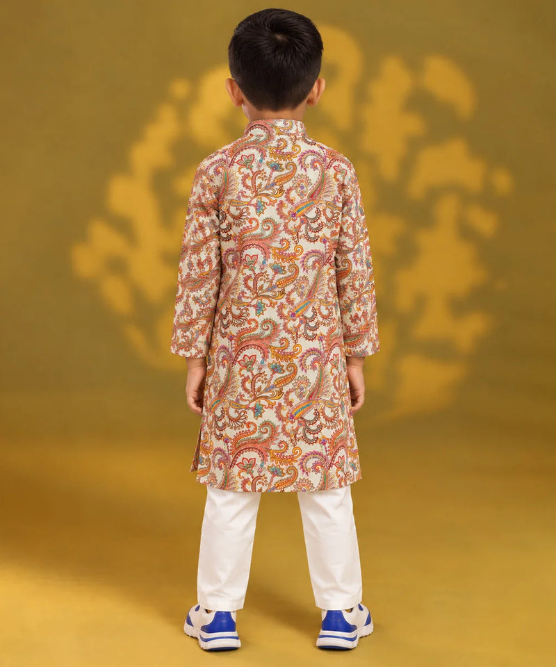 A child viewed from the back wearing a long-sleeve beige kurta with an intricate orange, red, and green paisley pattern, paired with white trousers and blue sneakers, set against a yellow background with a matching shadow.