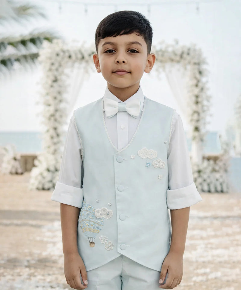 Young boy in formal attire with a light blue vest and white shirt against a decorative background.