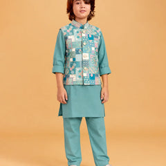 A child model wearing a teal green traditional Indian outfit with a patterned patchwork vest, standing against a solid beige background.