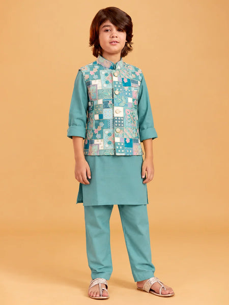 A child model wearing a teal green traditional Indian outfit with a patterned patchwork vest, standing against a solid beige background.