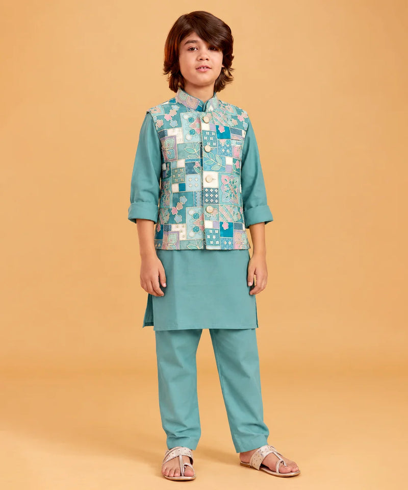 A child model wearing a teal green traditional Indian outfit with a patterned patchwork vest, standing against a solid beige background.